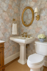 Neutral toned stone patterned wallpaper with gold mirror and sconce, featuring sink and toilet.
