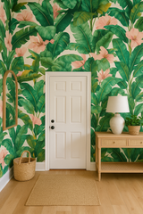 Palm Mirage Wallpaper with a green leaf and light pink flower pattern, against a white door, wooden table, and mirror.
