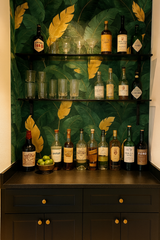 Jungle Luxe wallpaper with green leaves and gold accents, covering a bar setup with shelves of glassware and bottles.
