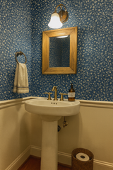 Blue floral wallpaper from Inkwash Brambletrail, covering the walls above white wainscoting in a bathroom with gold fixtures.
