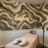 Abstract wallpaper with gold veining and gray marbling, featuring a neon "lashes" sign above a treatment bed.