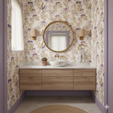 Violet Fête Wallpaper with floral and ribbon pattern, covering a bathroom wall with a mirror and vanity.
