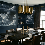 Dramatic cloudscape wallpaper by Nocturne covering a dining room wall with dark wood trim and gold chandelier.
