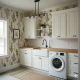 Botanical wallpaper in a laundry room with a green and cream pattern, beside a window and white cabinets.