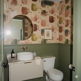 The wallpaper from Copper Mule Market features a pattern of colorful mugs and fruit slices, adorning a powder room with green wainscoting.
